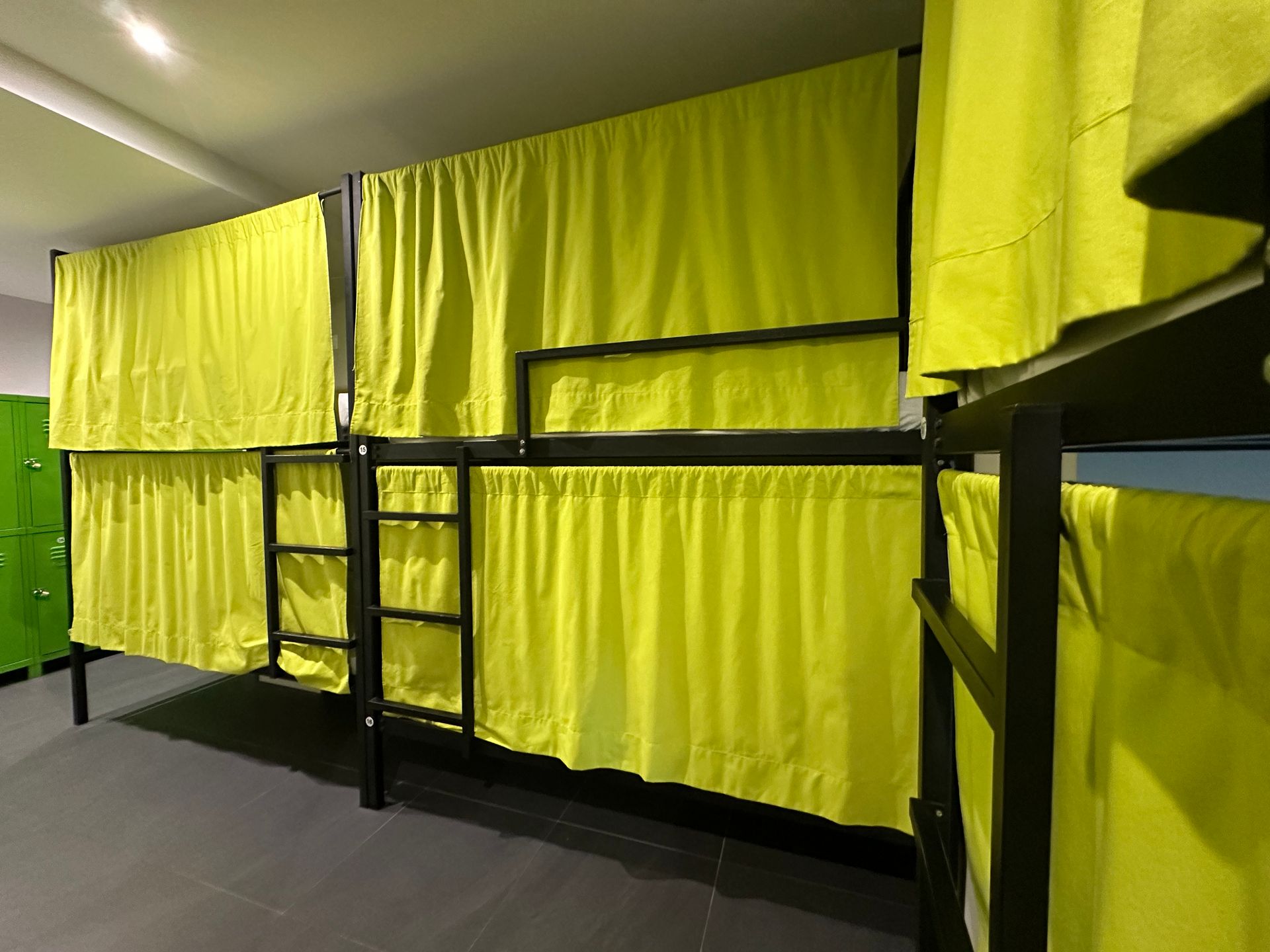 Literas con cortinas amarillas en una habitación de hostal. Estructuras metálicas oscuras y taquillas verdes.