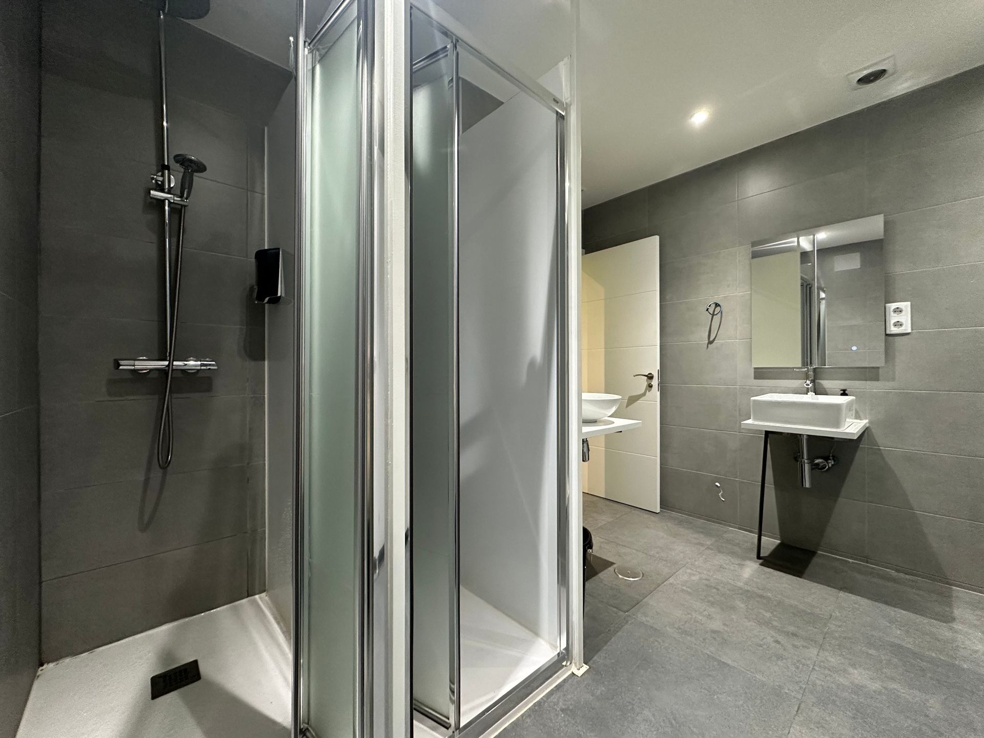 Baño moderno con paredes de azulejos grises, ducha, lavabo y espejo.