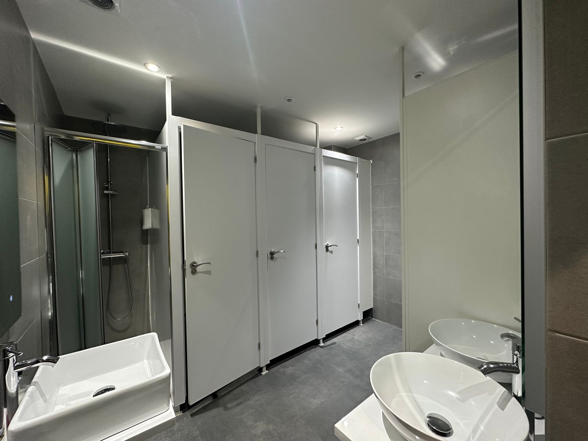 Baño público con cabinas blancas, lavabos y ducha. Paredes y suelo grises.