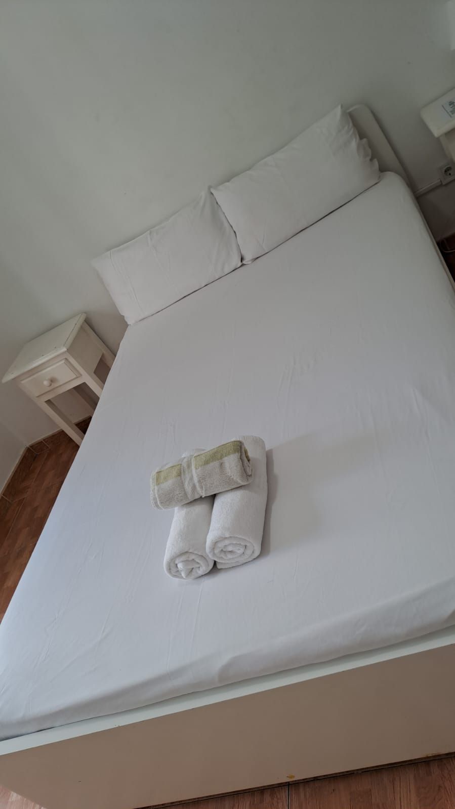 Dormitorio con cama blanca, dos almohadas, toallas enrolladas y mesita de noche.