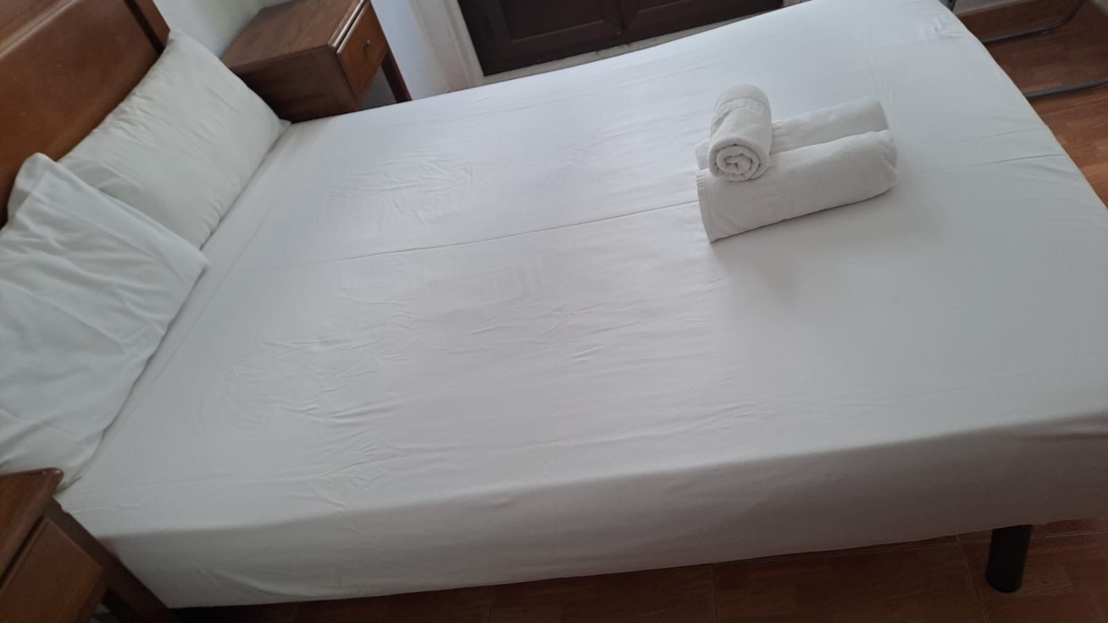 Cama con sábanas blancas y toallas enrolladas encima, cerca de una mesita de noche.