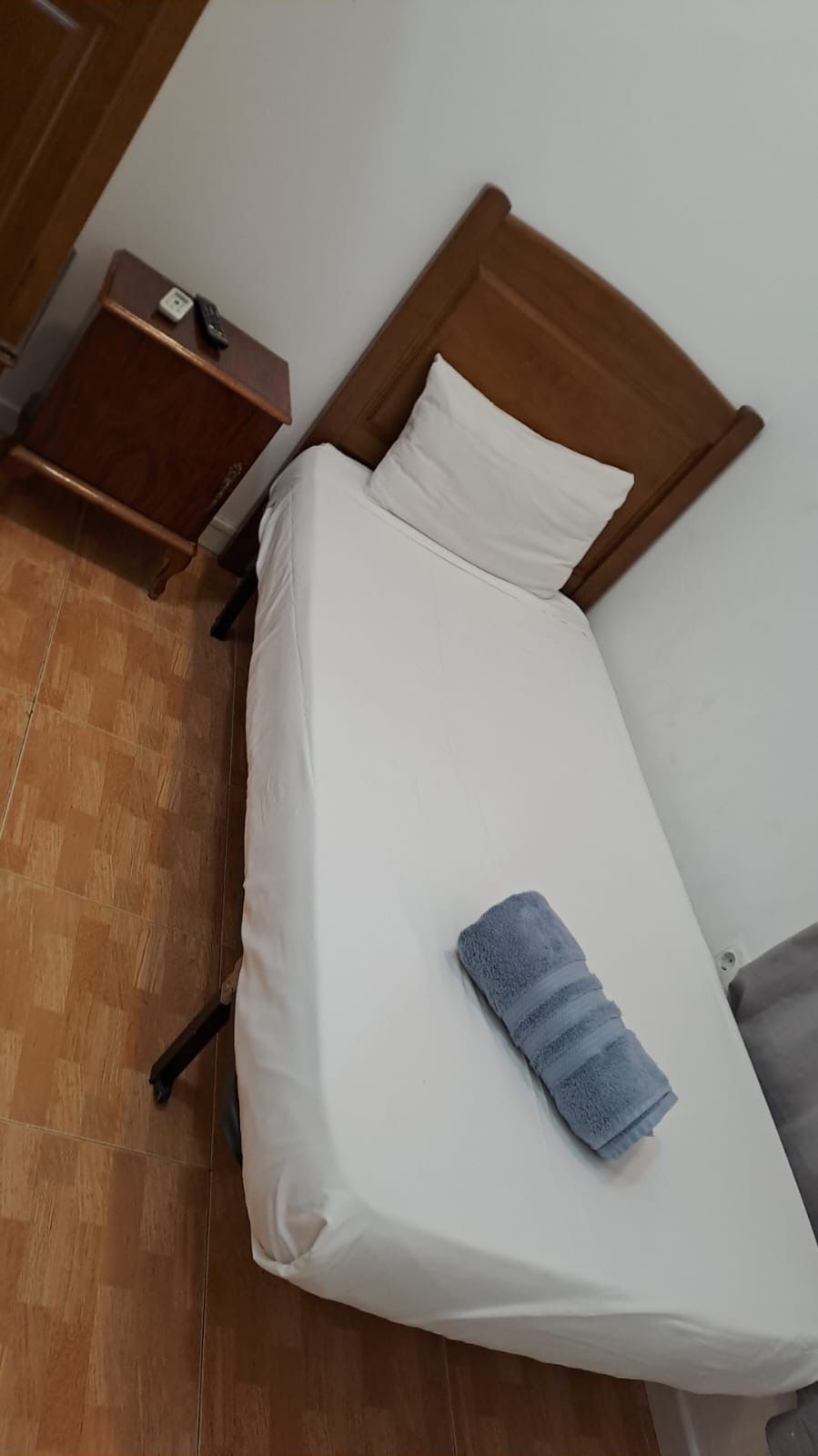 Una cama individual con sábana blanca, almohada y toalla azul. Junto a ella hay una mesita de noche.