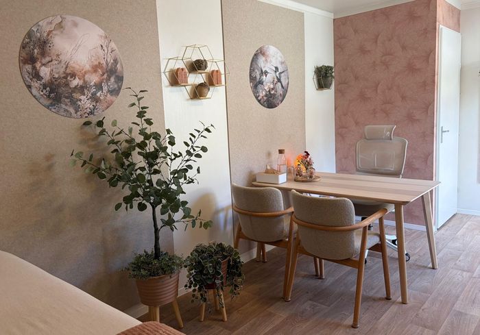 Gezellige praktijkruimte met houten tafel, twee stoelen, roze bloemenmuur en een kamerplant bij het raam.