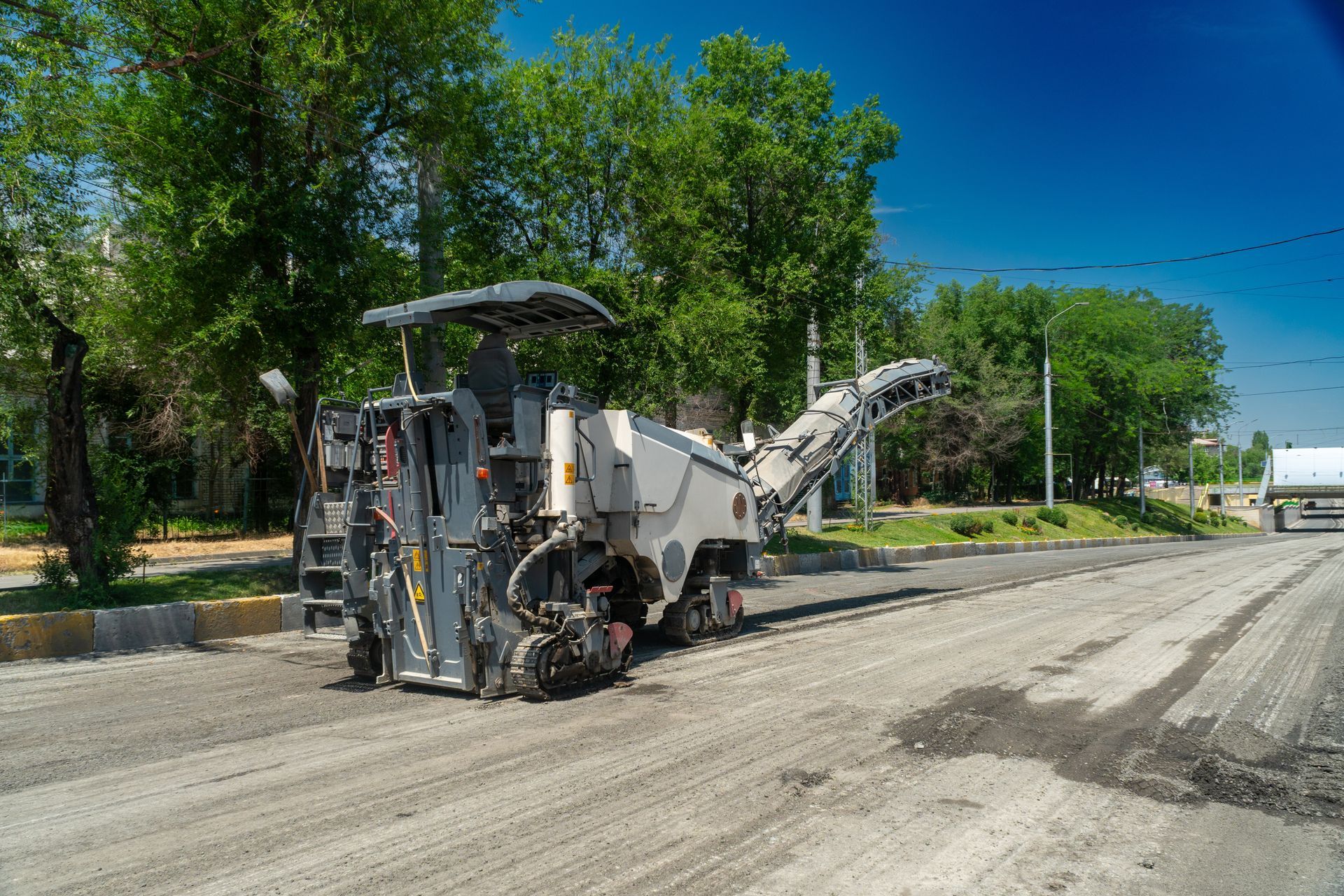 Road milling machine on a street, removing asphalt. Trees and blue sky in the background.