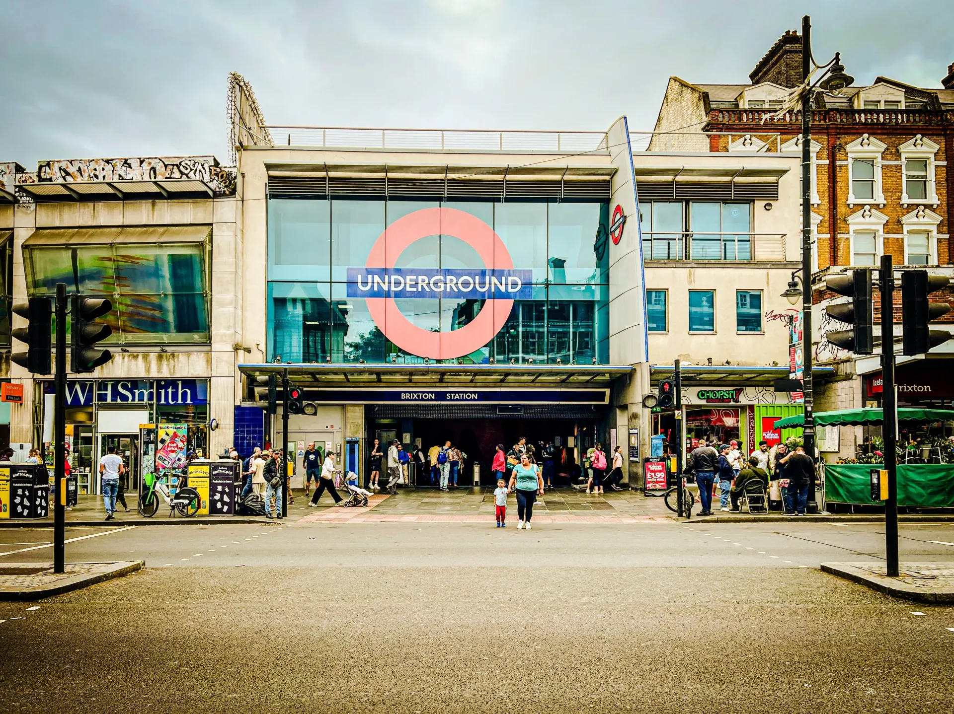 Underground station entrance with red roundel sign, people walking outside, and historic buildings nearby