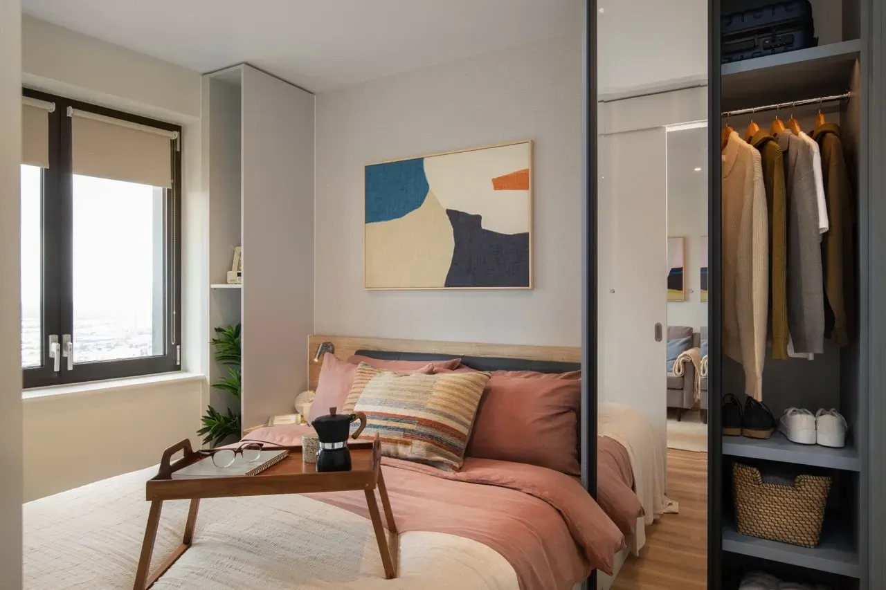 A modern bedroom with a bed featuring pink linens, a breakfast tray, abstract wall art, and an open wardrobe.
