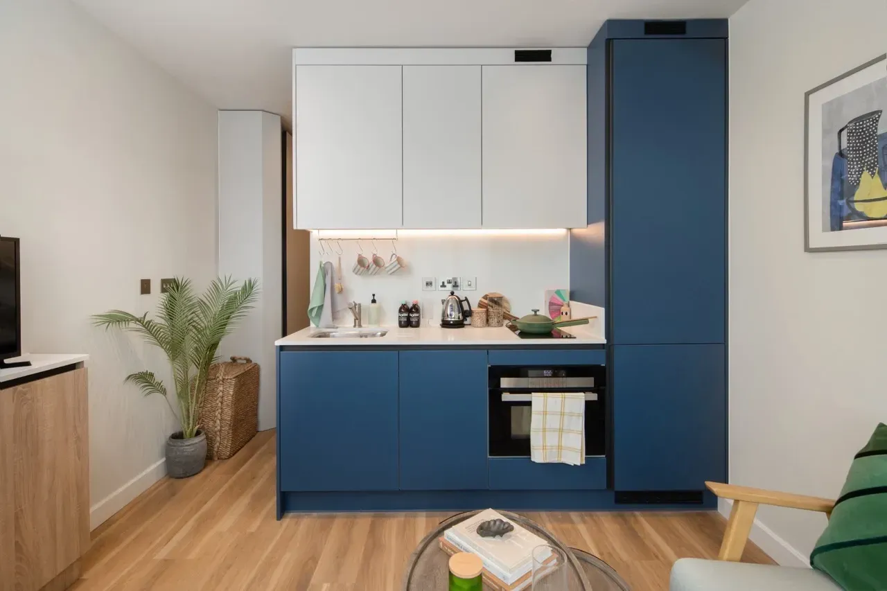 A modern kitchen with blue lower cabinets, white upper cabinets, a built-in oven, and a tall pantry on a wooden floor.