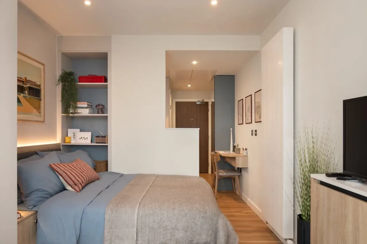 A modern, well-lit studio apartment featuring a bed with blue bedding, a side shelf, a small desk, and a television.