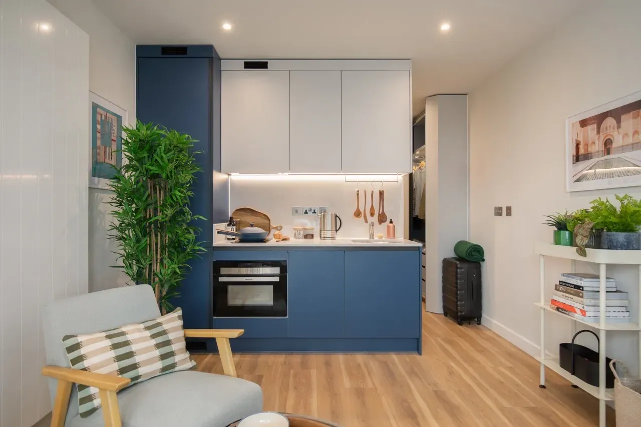 A modern kitchenette with blue and white cabinets, an oven, and a green plant, next to a light-colored armchair.