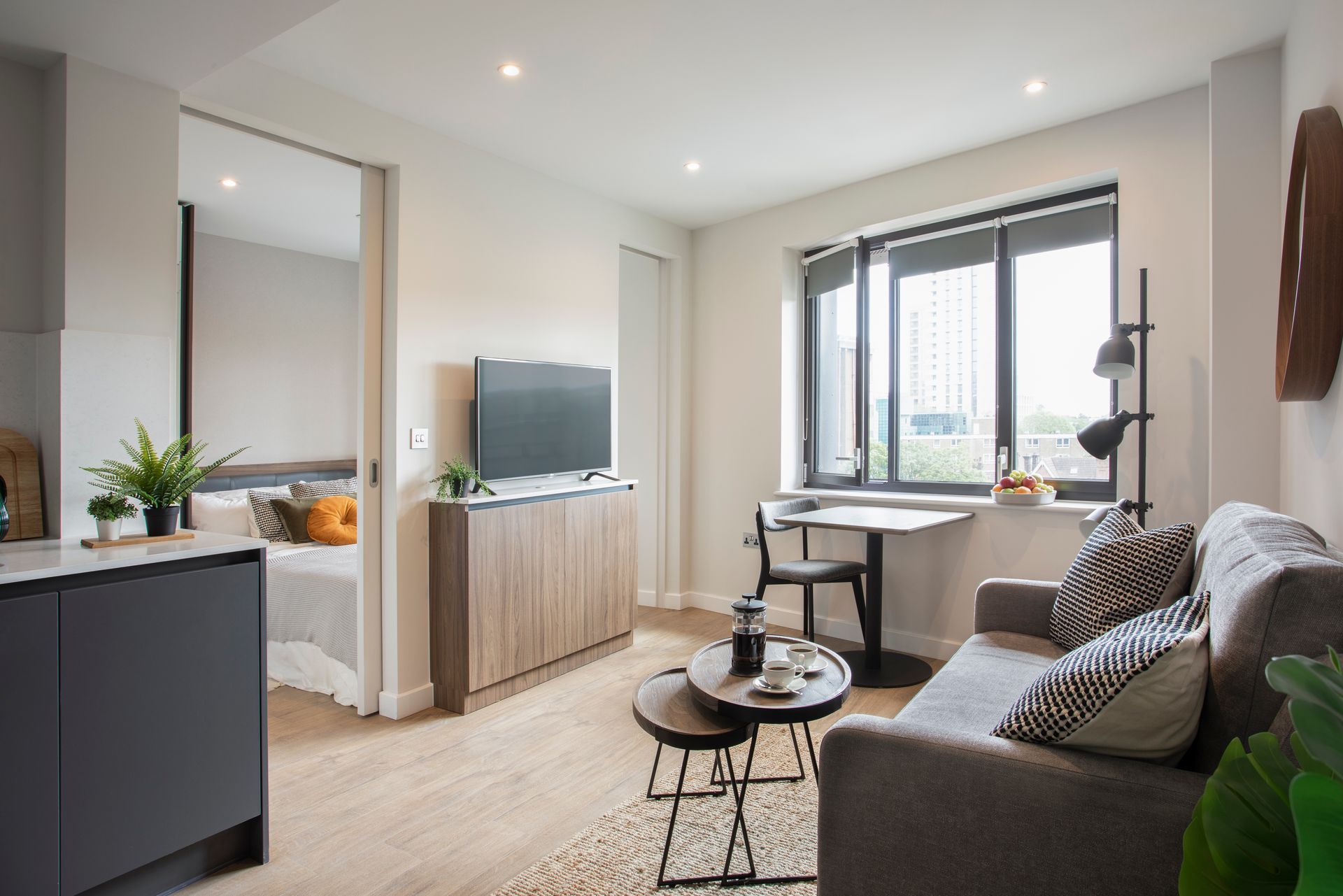 A bright, modern studio apartment featuring a grey sofa, a small dining table, a TV on a wooden console, and a bedroom area.