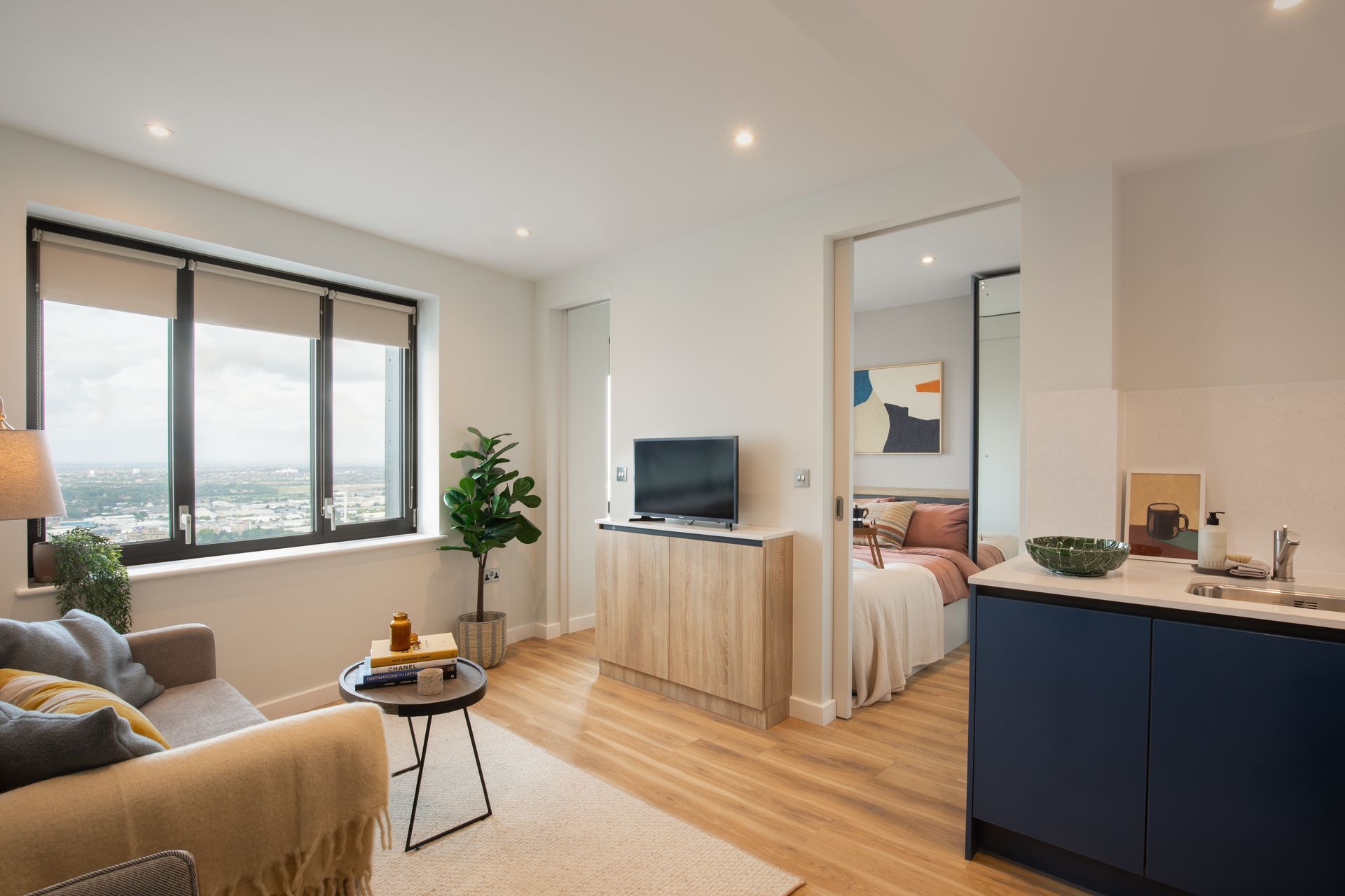 A modern apartment living room with a sofa, TV console, and view of a bedroom through an open doorway.