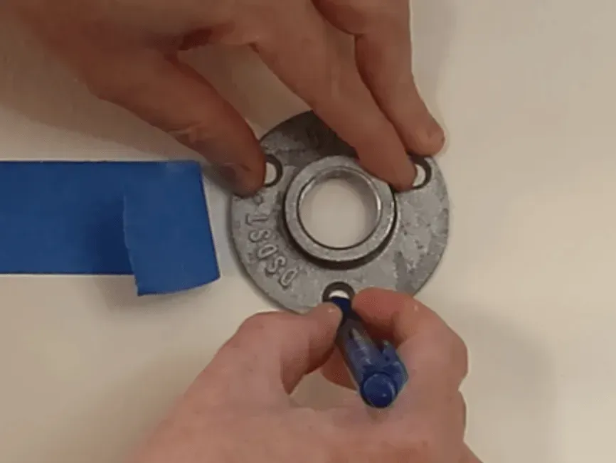 Hands using a pen to mark drill holes through a metal floor flange onto a white surface next to blue painter's tape.