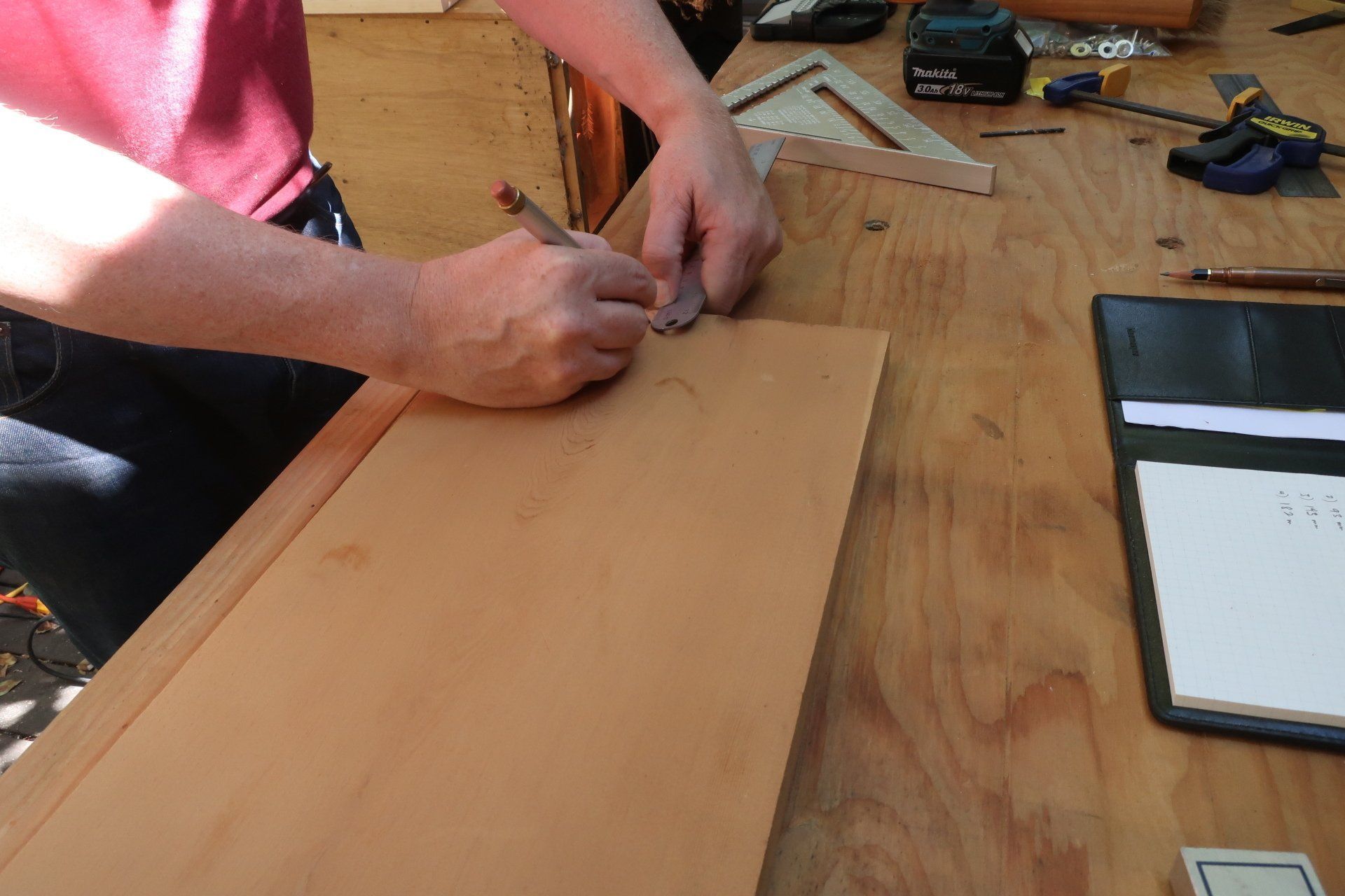 Craftsperson using a pencil and a metal guide to mark a wooden panel on a busy workshop table.