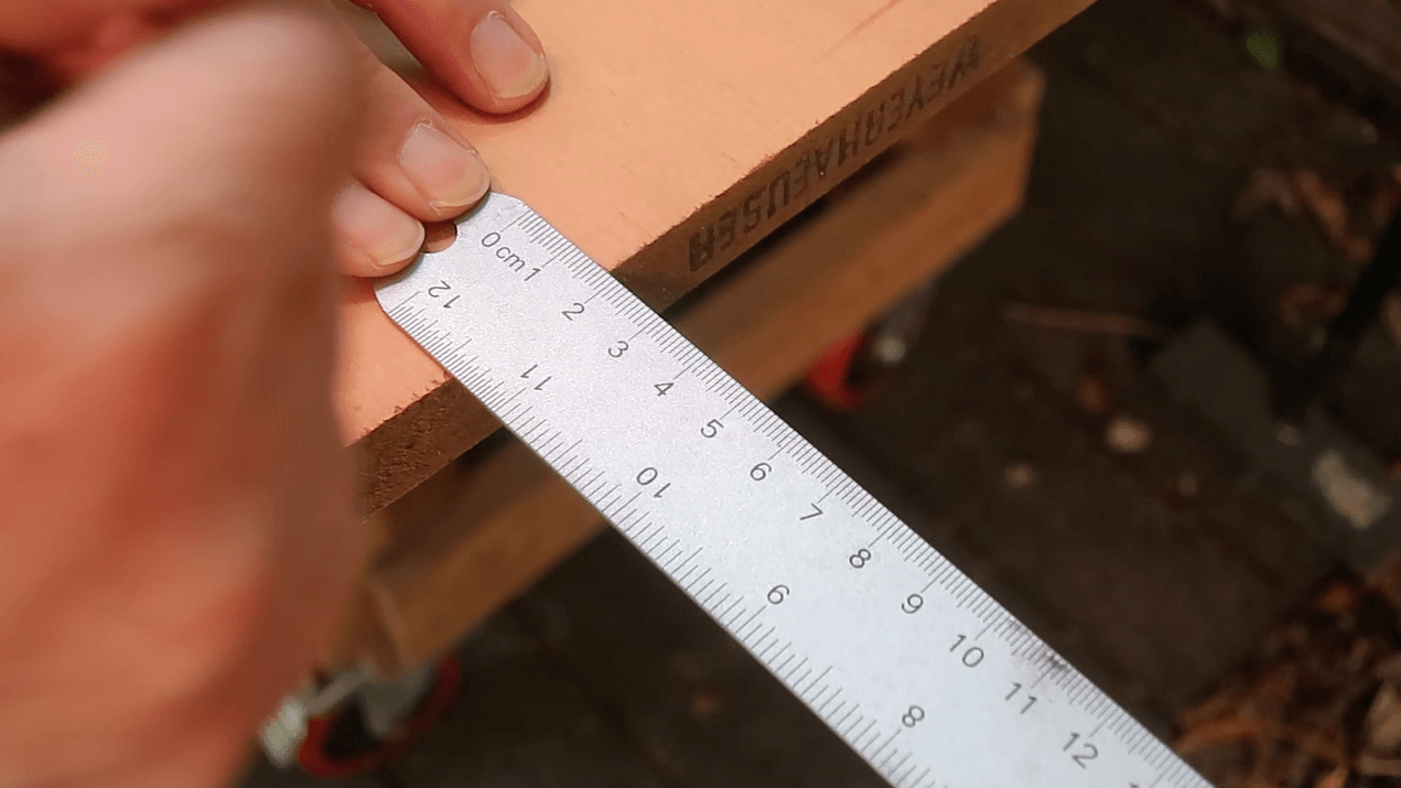 A high-angle, close-up shot of a hand using a metal ruler to measure exact points on a light wood panel for a cat shelf.