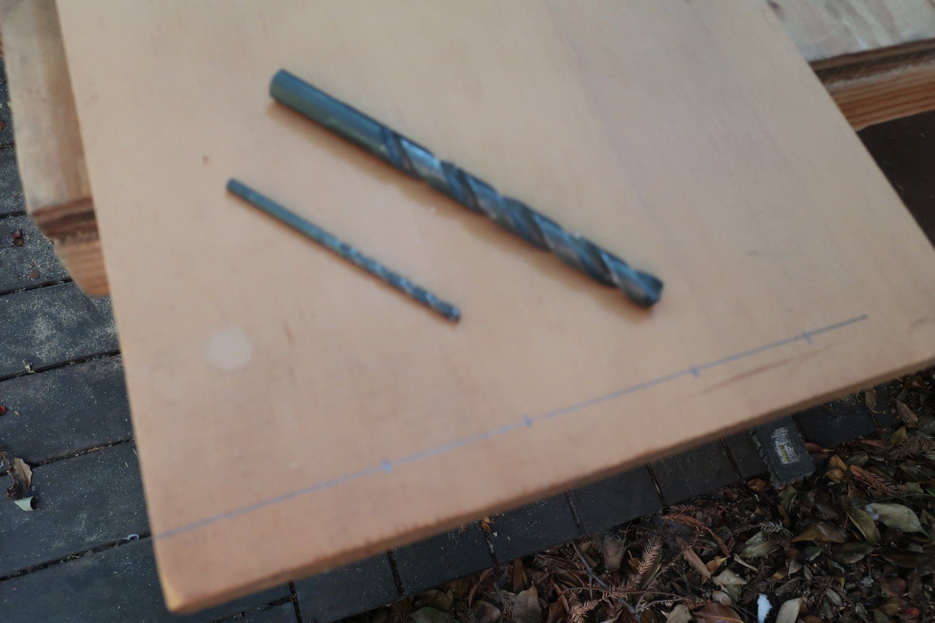 Two different-sized metal drill bits resting on a light-colored wooden panel marked with a pencil line and guide points.