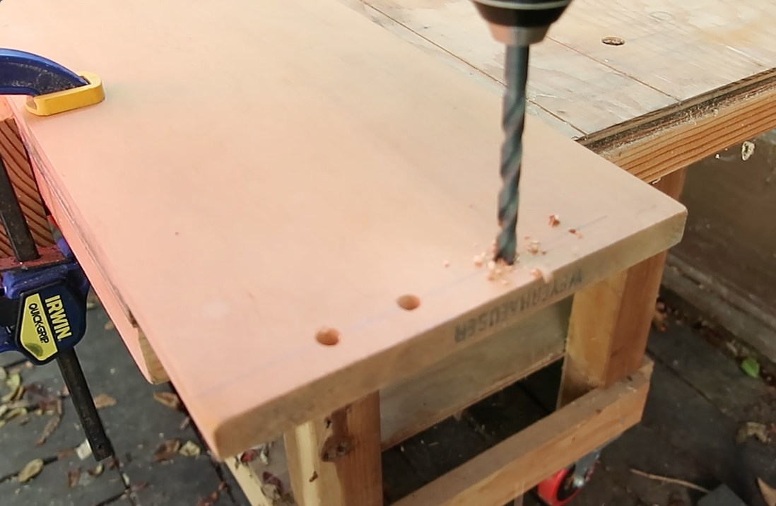 Action shot of a power drill bit creating a precise hole in a light-colored wooden shelf component clamped to a workbench.