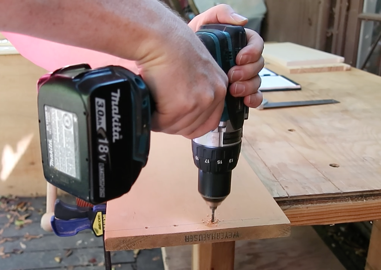 Close-up of a person using a cordless Makita power drill to create pilot holes in a wooden shelf panel on a workshop table.