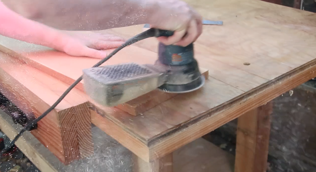 Action shot of a person using a power sander on a piece of light-colored wood.
