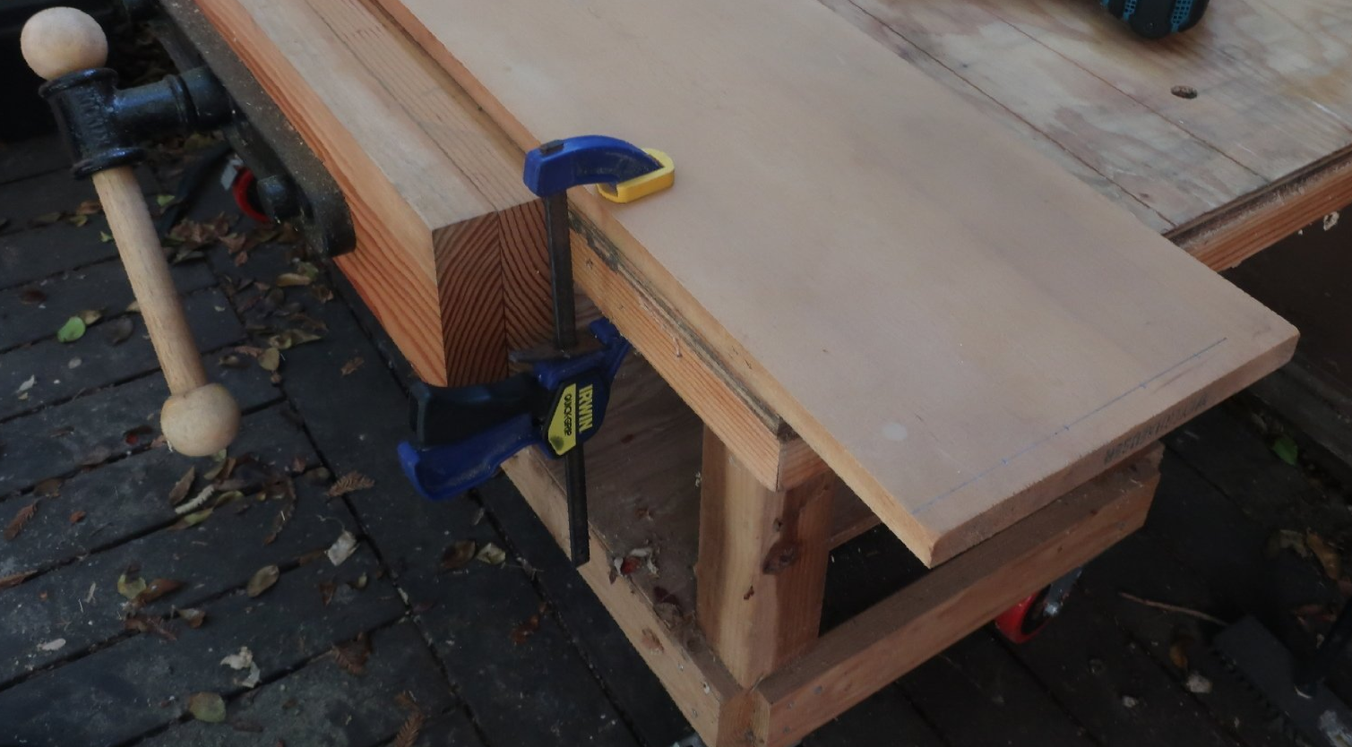 A wooden shelf panel securely held to a heavy-duty workbench using a blue and yellow Irwin bar clamp.