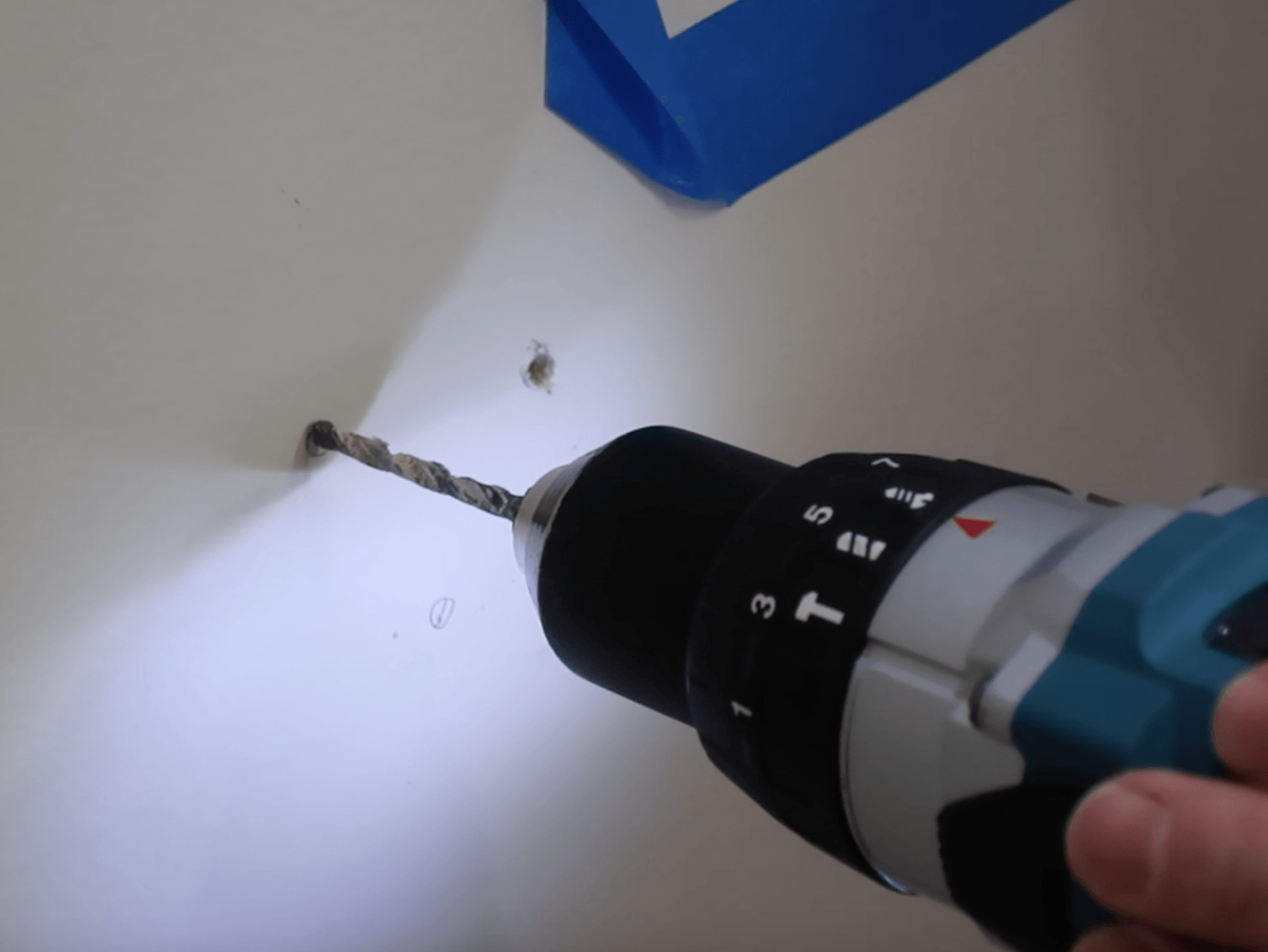 Close-up of a power drill boring a hole into a white wall near blue painter's tape for mounting hardware.
