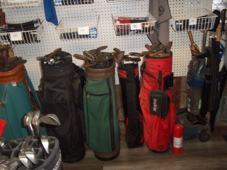 Several golf bags in various colors, including black, green, and red, stand in a row with clubs inside against a pegboard.