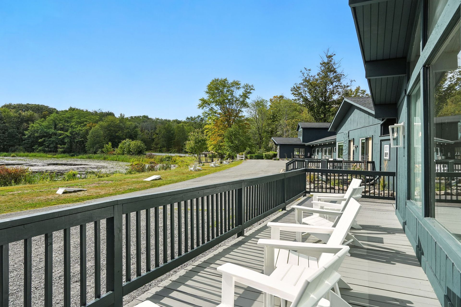 A wooden deck with white chairs overlooking a gravel path and trees under a blue sky.