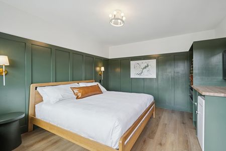 Green-paneled hotel bedroom with a wooden bed, white bedding, brown pillow, and light-colored flooring.