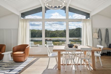 Spacious dining room with a large window overlooking a lake and trees; light wood table and chairs.
