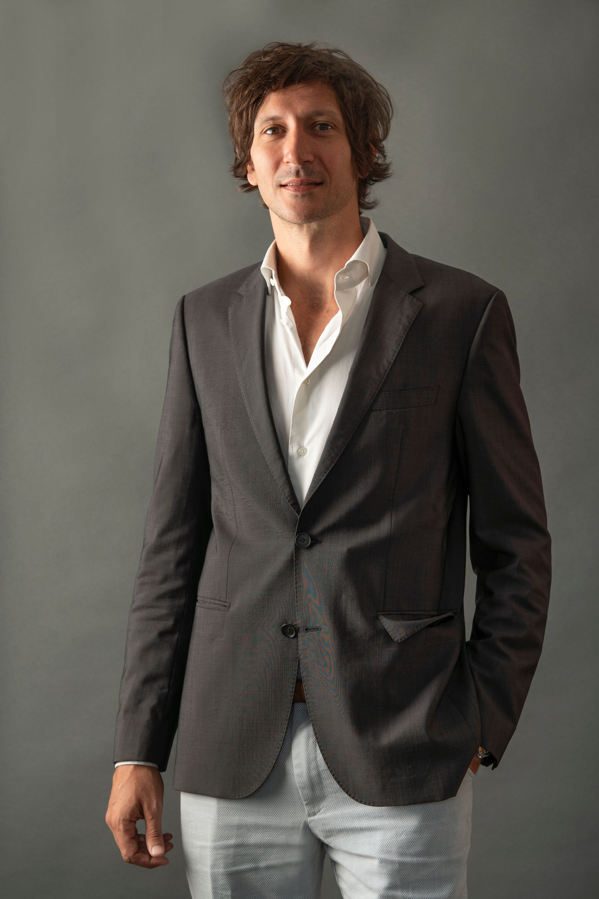 Un uomo con una giacca scura e una camicia bianca aperta, in piedi con una mano in tasca, su uno sfondo grigio.