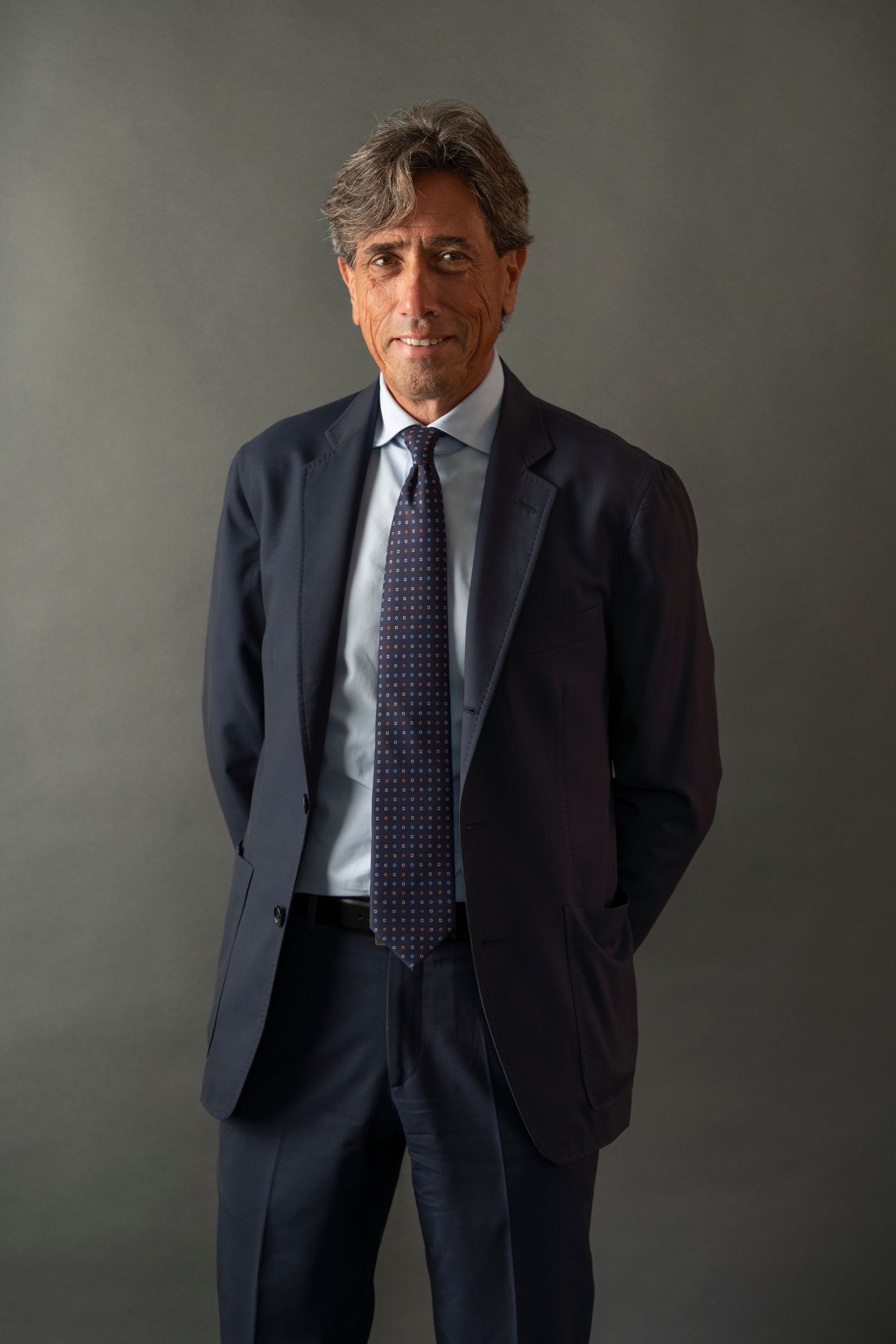 Uomo in abito blu navy, camicia azzurra e cravatta fantasia, in piedi con le mani dietro la schiena su uno sfondo grigio.