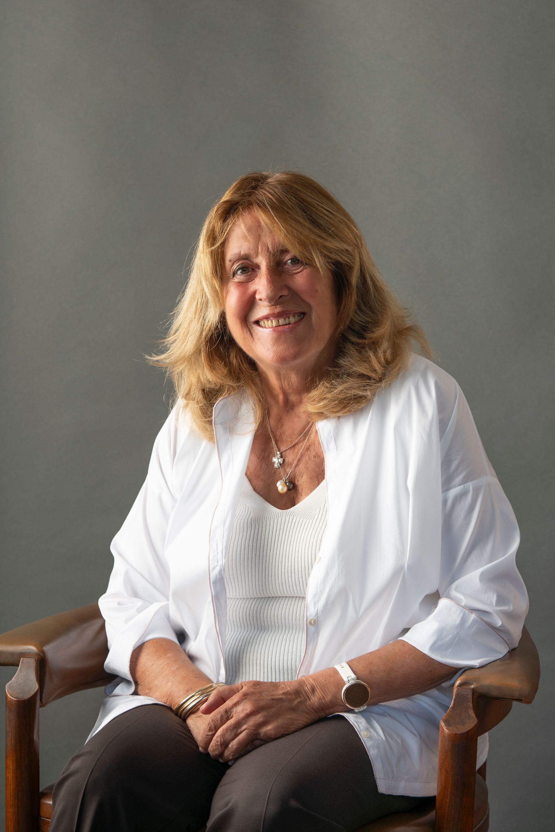 Donna sorridente con i capelli biondi, che indossa una camicia bianca, seduta su una sedia di legno, su uno sfondo grigio.