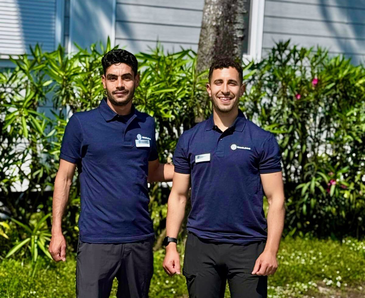 Due uomini in polo blu, sorridenti, in piedi all'aperto vicino al fogliame verde.