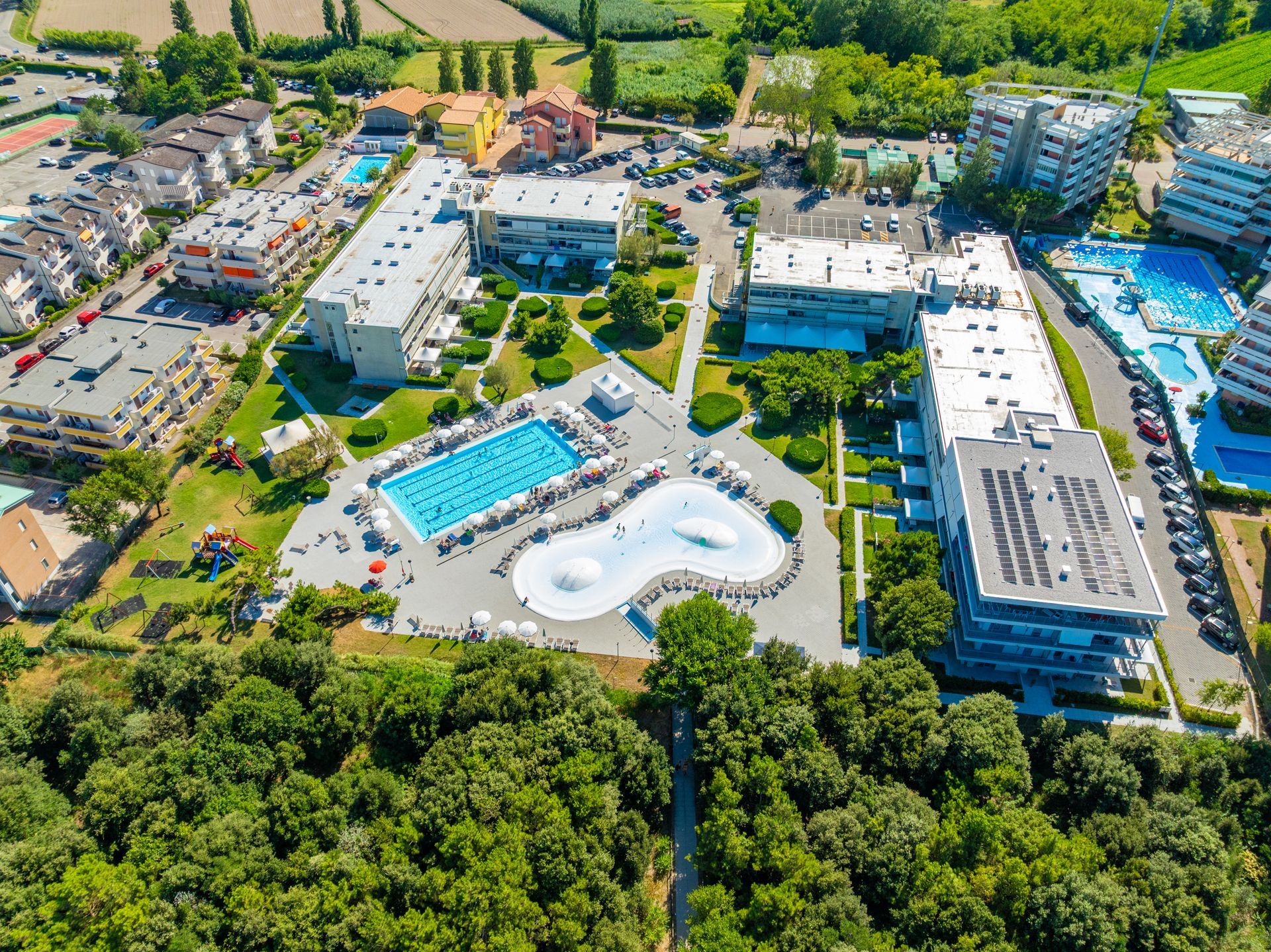 Vista aerea del resort con piscine, edifici e alberi circostanti in un ambiente soleggiato.