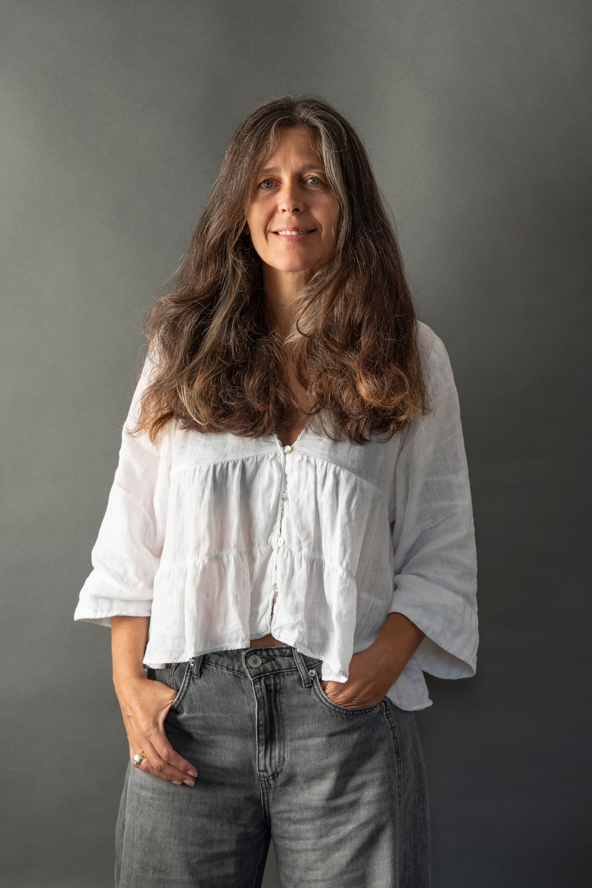 Woman with long wavy hair, wearing a white shirt and grey pants, hands in pockets, smiling, against a grey backdrop.