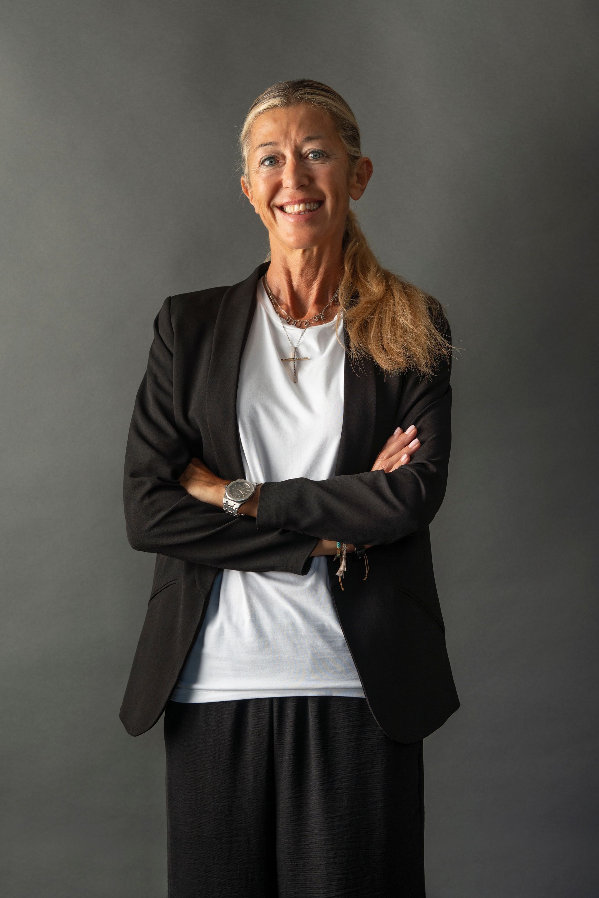 Donna con i capelli biondi, giacca e pantaloni neri, braccia incrociate, sorridente, su uno sfondo grigio.