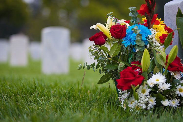 Colorful bouquet of red, yellow, and white flowers on grass in a cemetery with blurred gravestones in the background