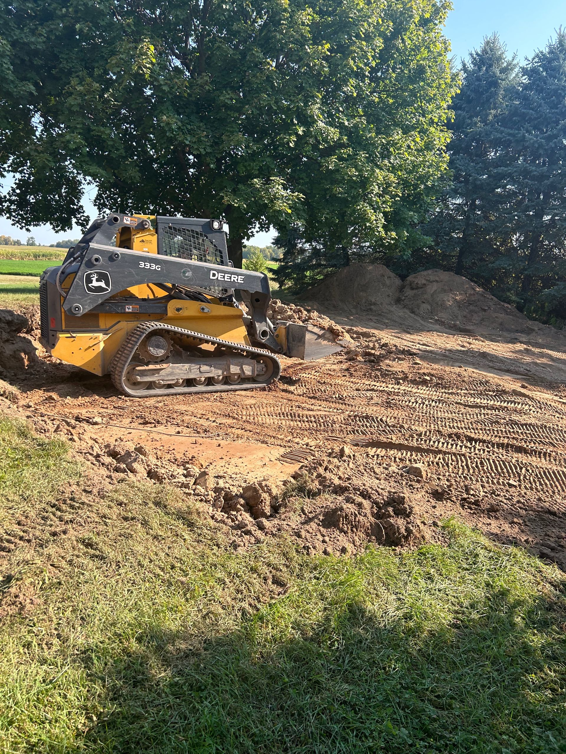 Yellow tracked skid steer moving dirt near a tree and soil piles in a field