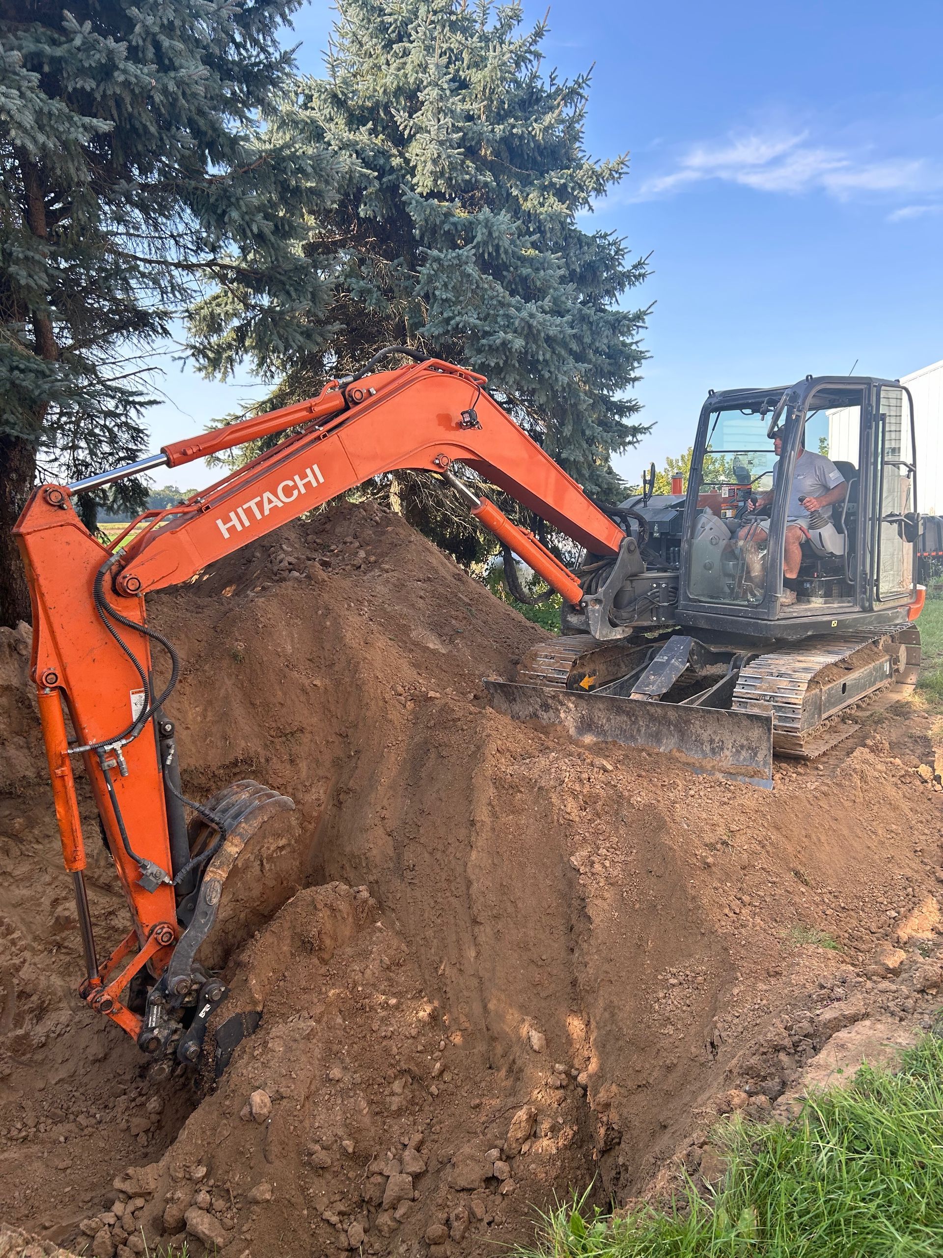 Orange excavator digging a large dirt mound beside a tree on a wooded work site.