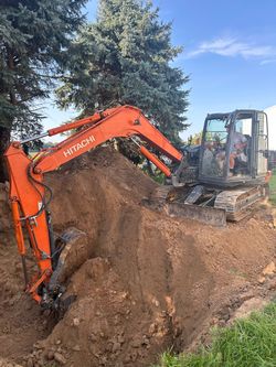 Orange excavator digging a large dirt mound beside a tree on a wooded work site.