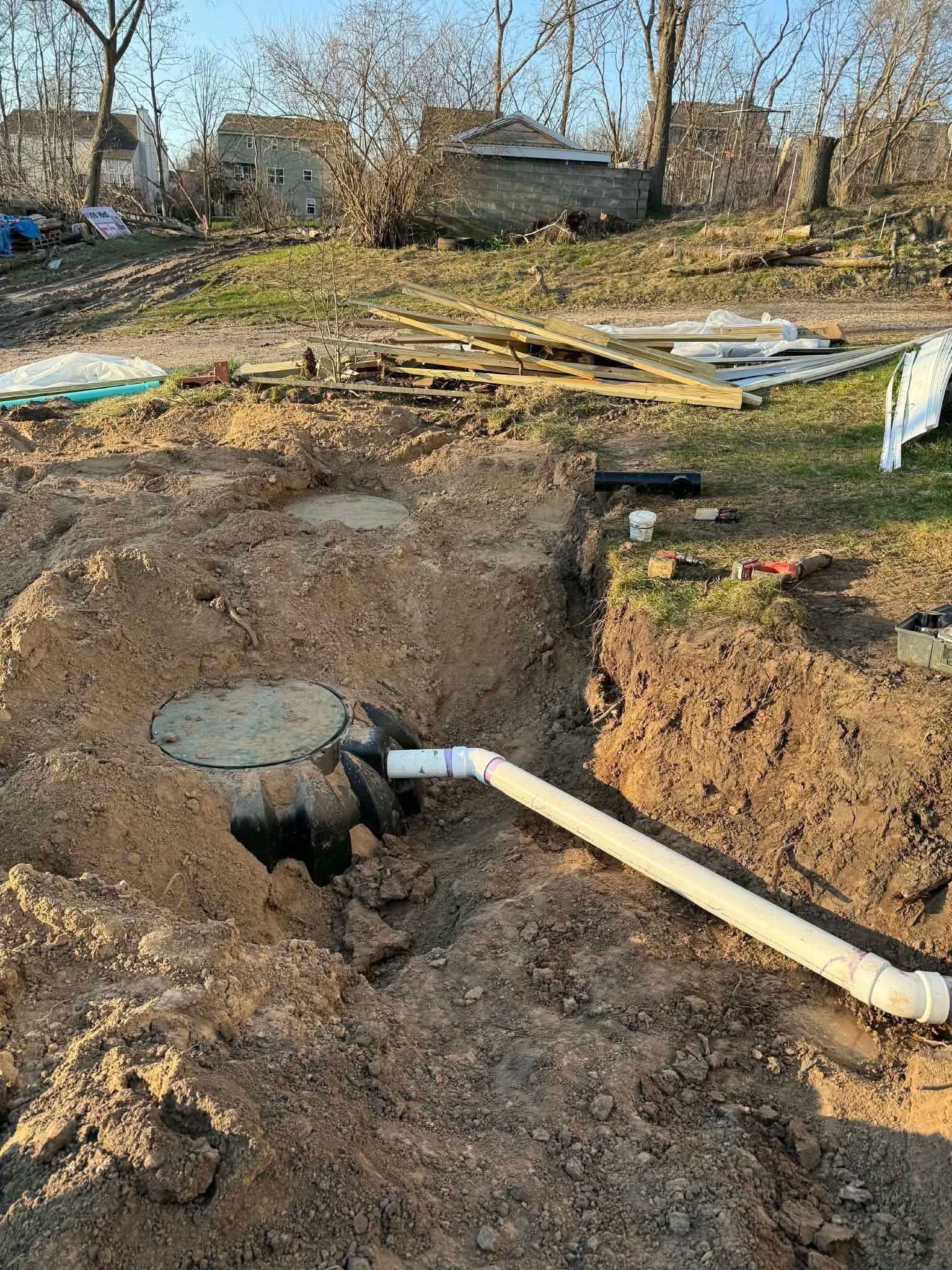 Damaged yard with a broken sewer pipe and exposed utility hole in muddy ground, trees and debris in background