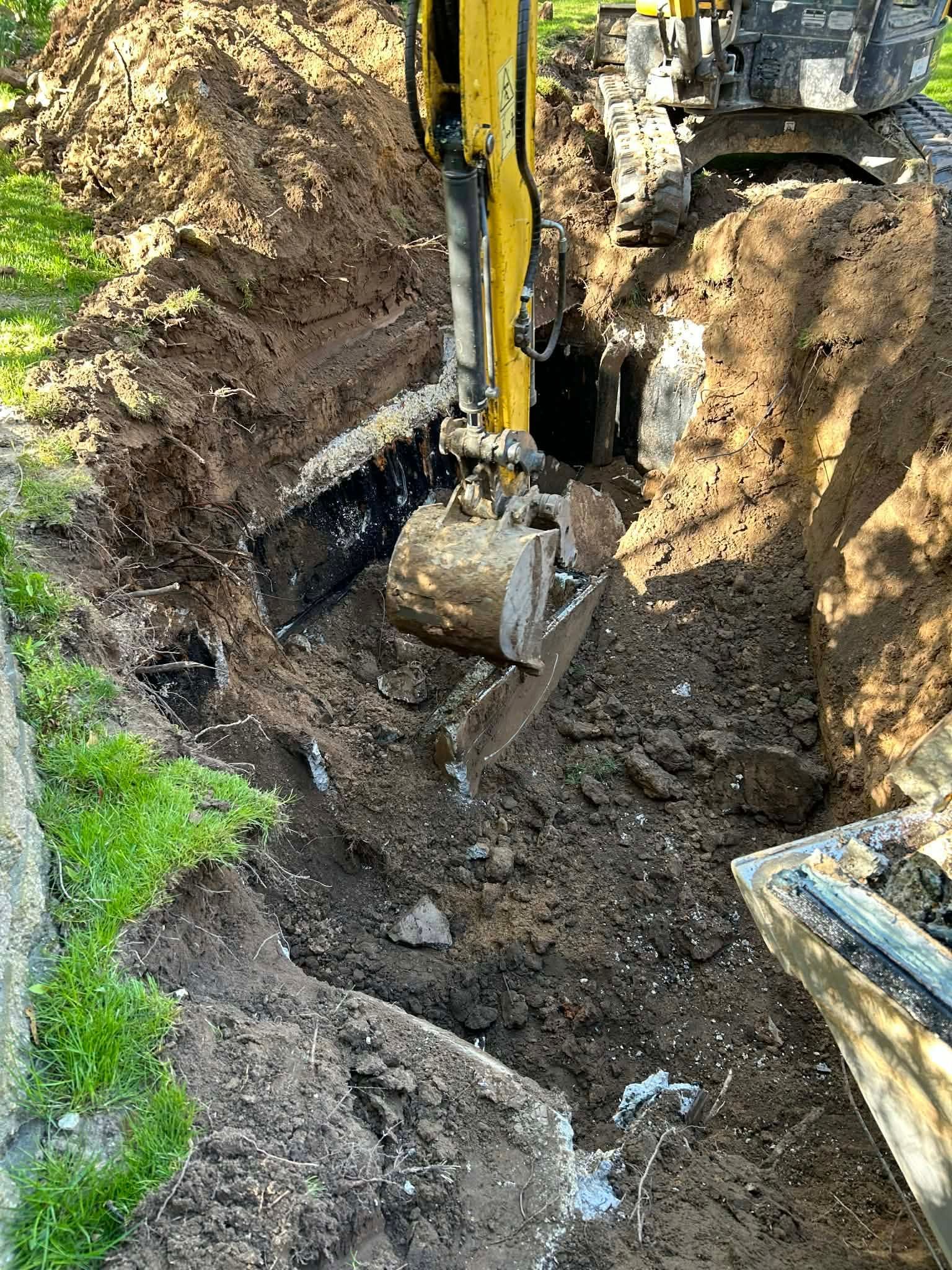 Excavator digging a trench beside a path, with muddy soil and a grassy edge.
