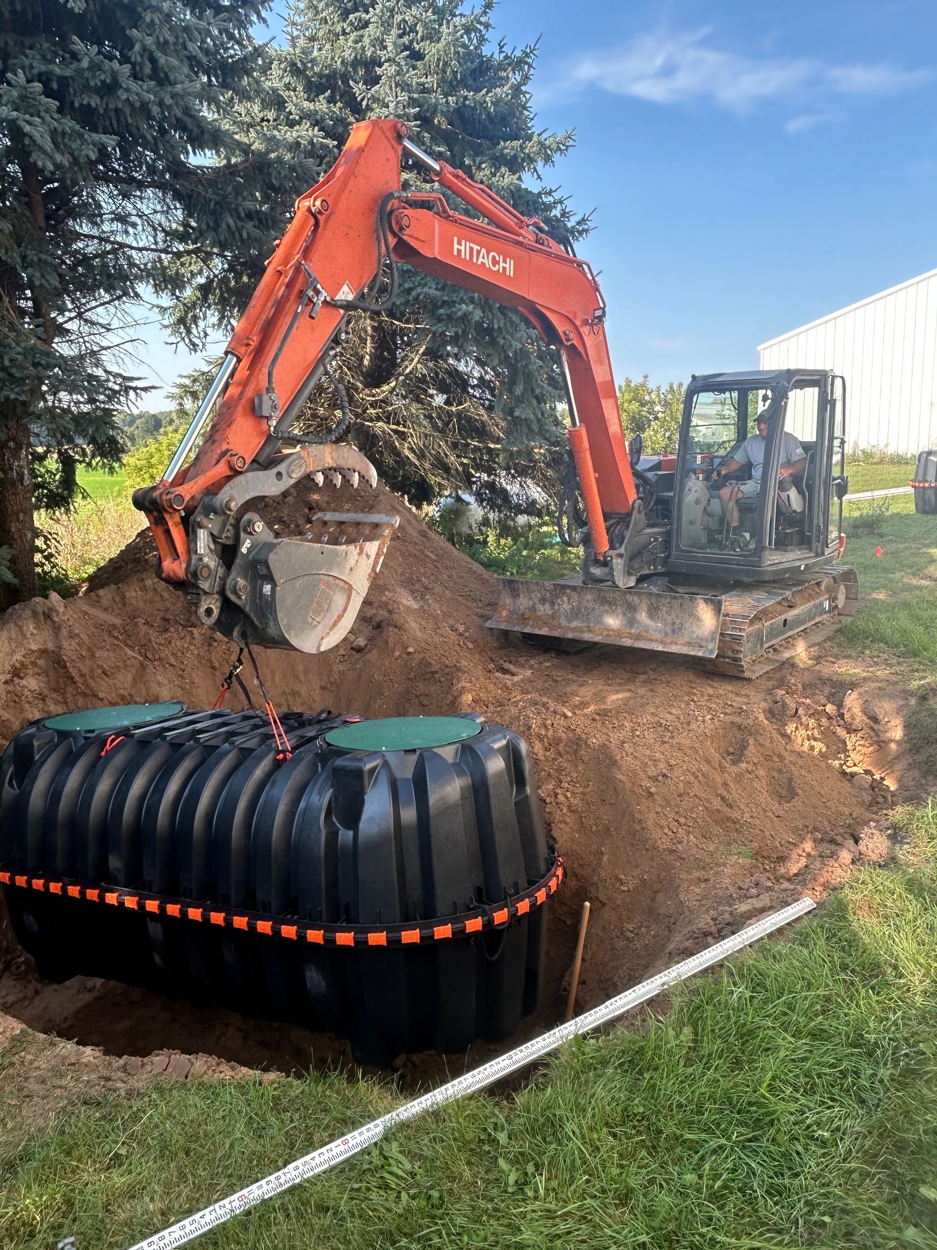 Excavator digging soil beside a black plastic tank in a grassy yard under a blue sky