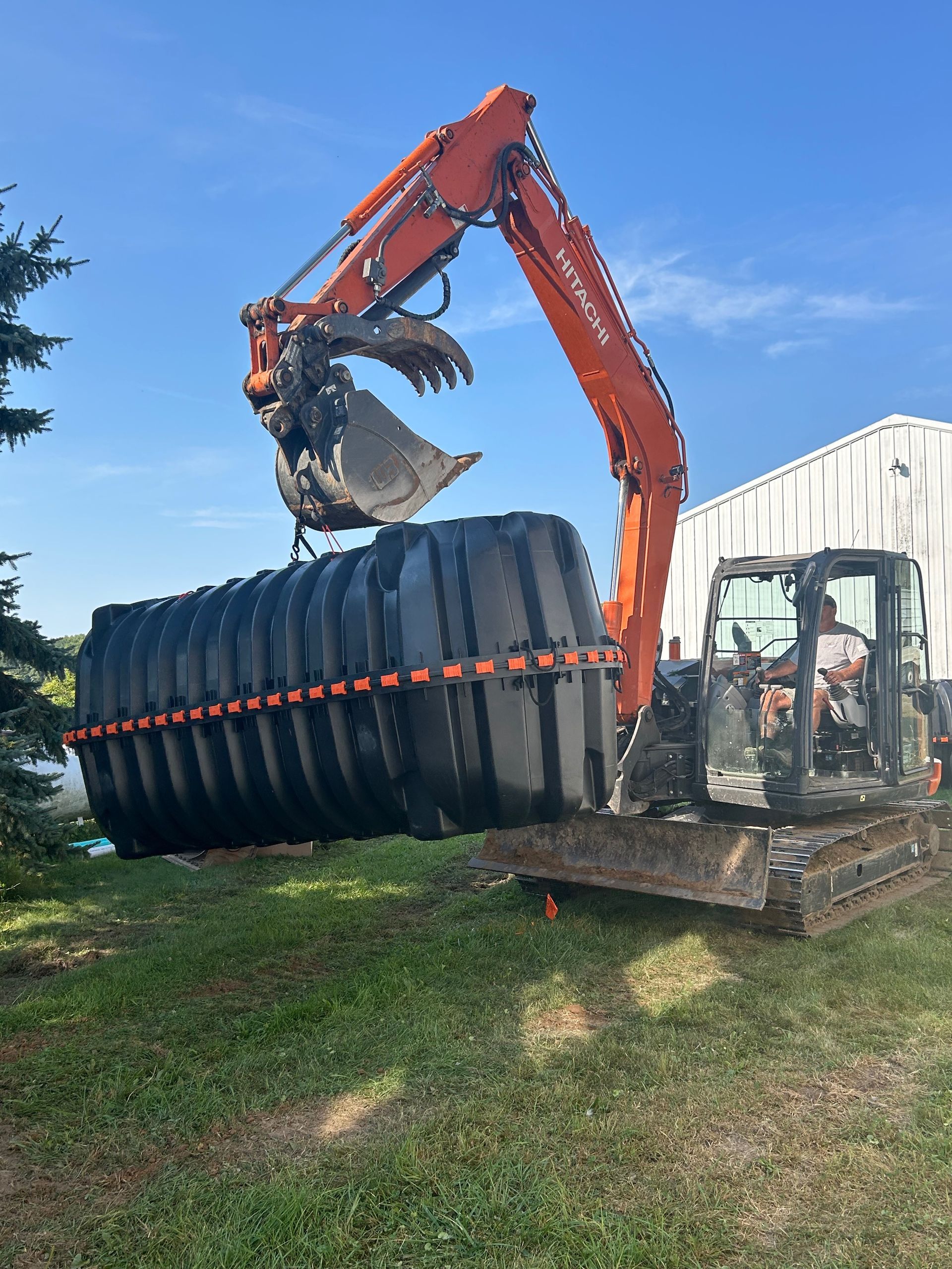 Orange excavator lifting a large black tank on a grassy lot under a blue sky