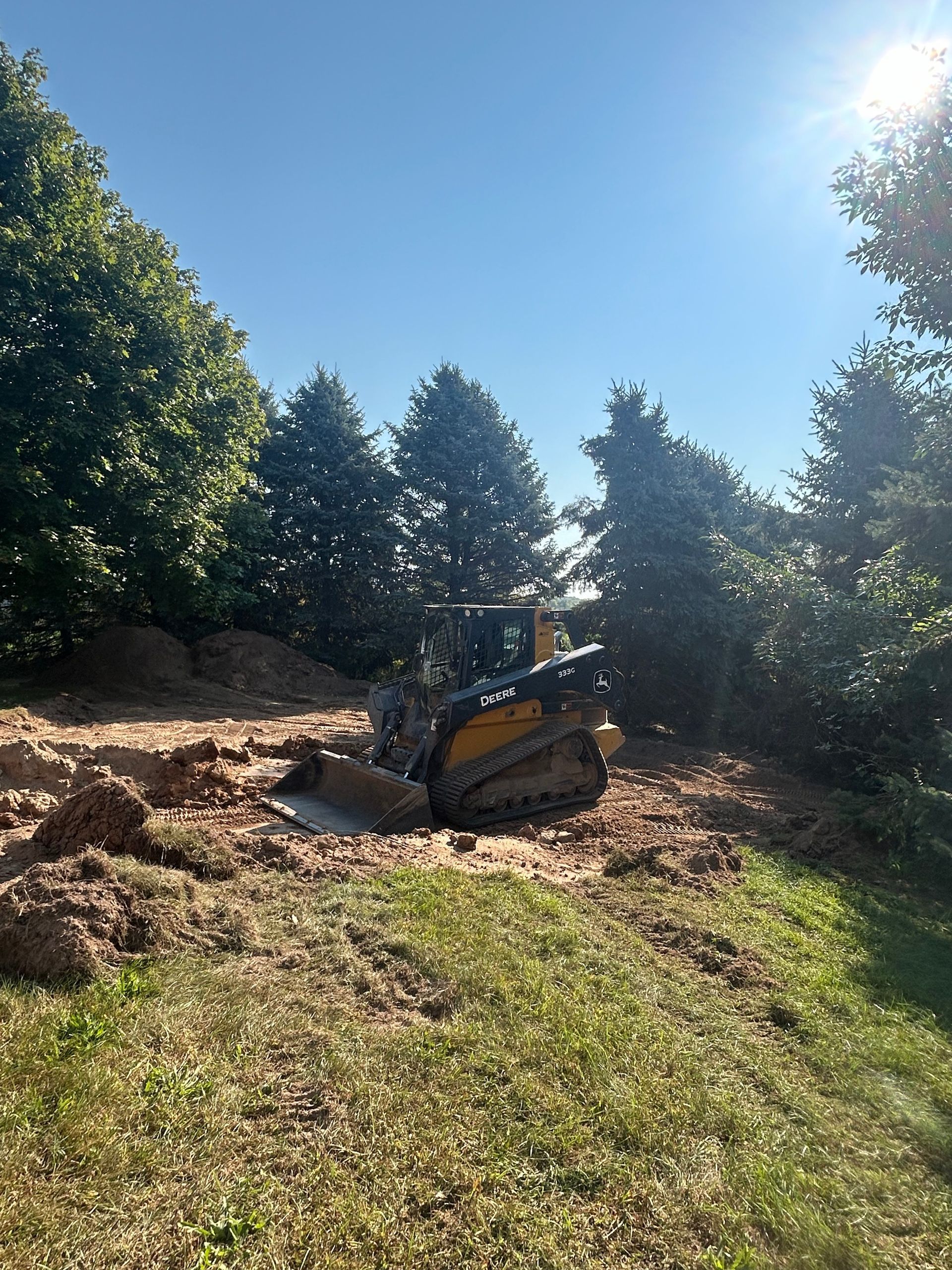 Small bulldozer on a grassy dirt slope under bright sun, surrounded by trees and blue sky