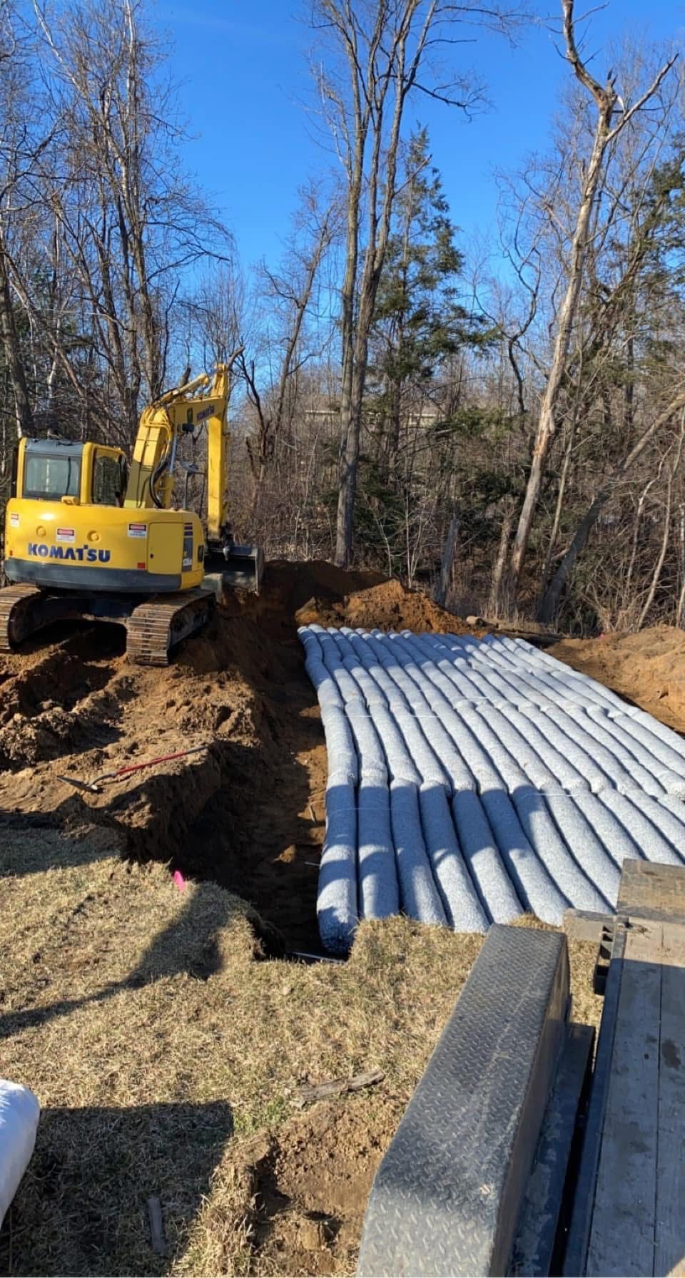 Yellow excavator beside stacked corrugated metal pipes on a dirt construction site in a wooded area