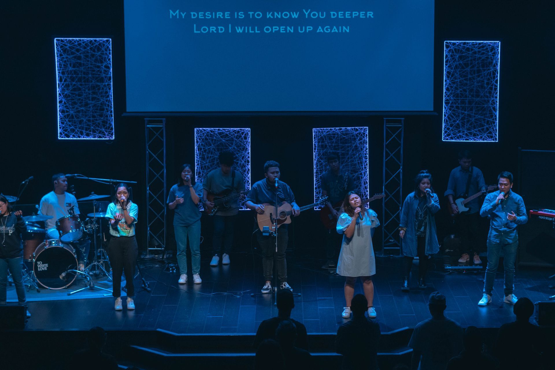 A worship band performing on stage under blue lights; singers, musicians, and lyrics on a screen.