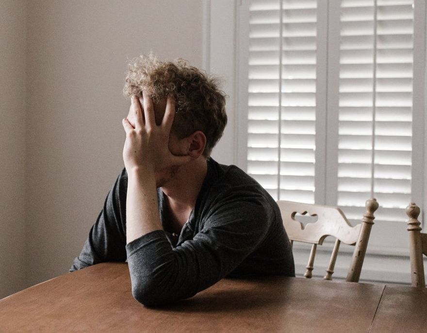 Person with curly hair, covering face with hand at wooden table, near window with closed blinds.