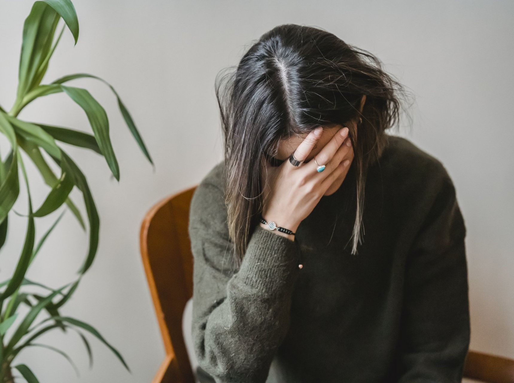 Woman with hand covering face, sitting in a wooden chair near a plant, looking distressed.