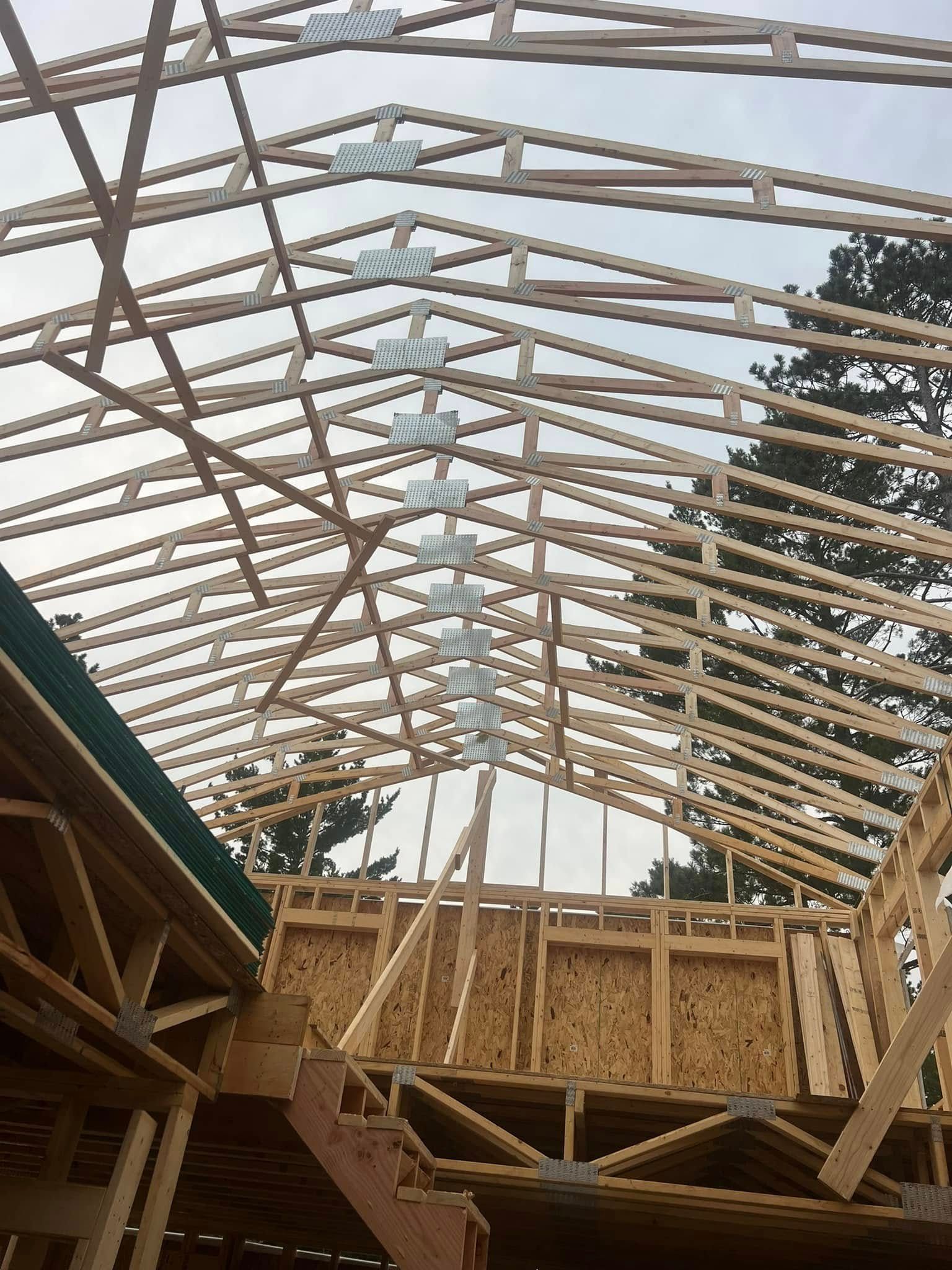Wood roof framing under construction, with exposed trusses and plywood walls against a cloudy sky.