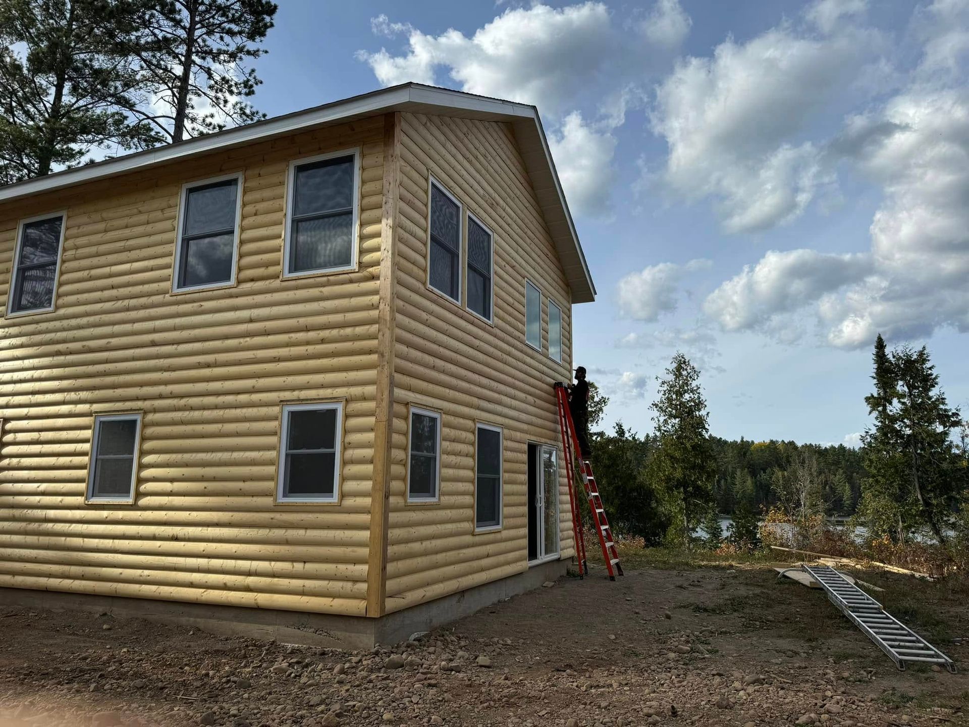 Two-story tan house with ladder leaning against it, set on a wooded lot under a cloudy sky